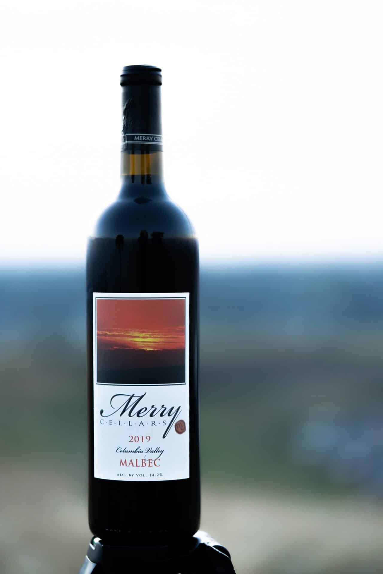 A bottle of Merry Cellars' 2019 Malbec Wine with a blurred outdoors backdrop. This is a red wine.